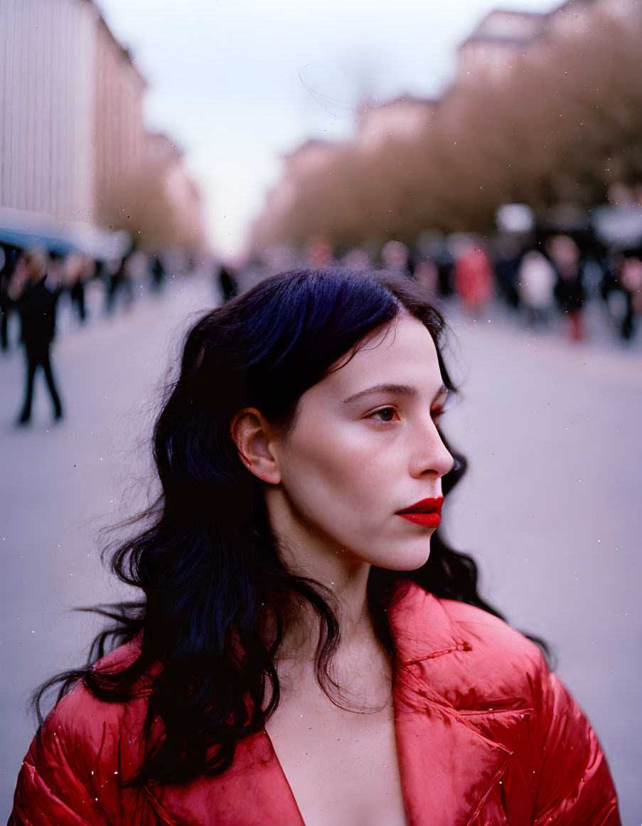 Dark-haired woman in red lipstick and jacket on tree-lined street.