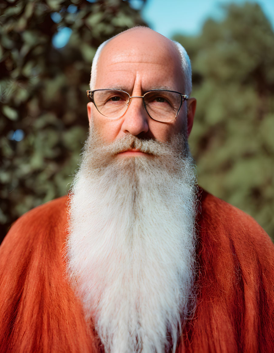 Elder man in white beard and orange robe with glasses in front of green foliage