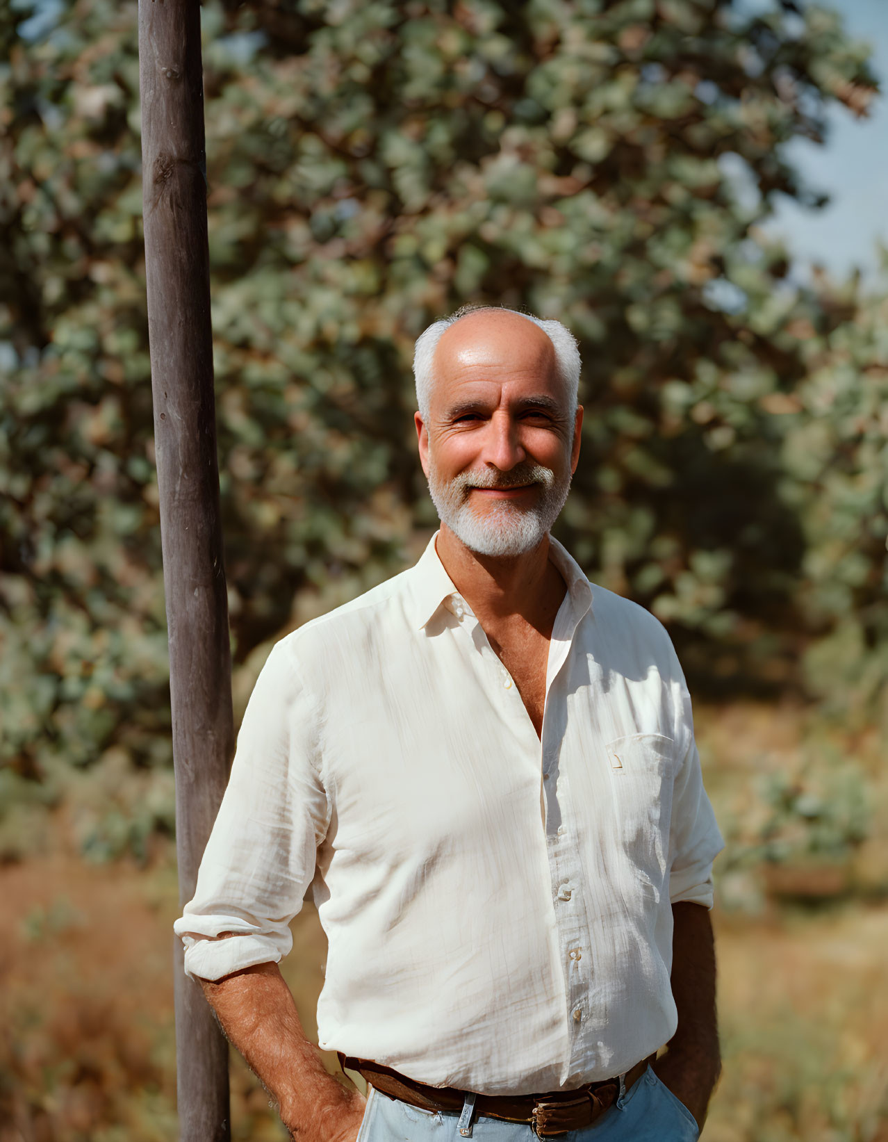 Elderly man with white beard and mustache outdoors among trees.