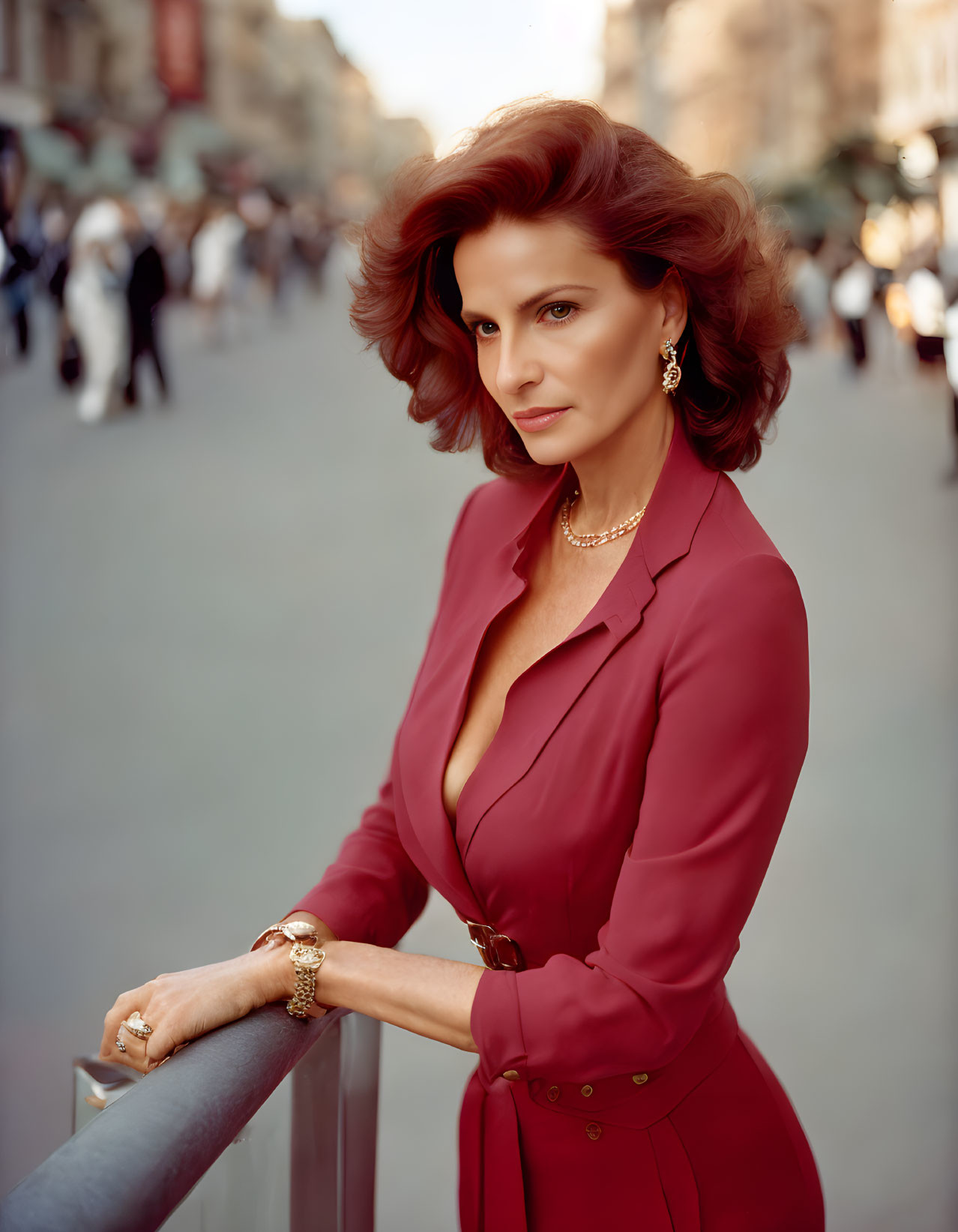 Auburn-haired woman in red blouse leaning against railing with blurred pedestrians.