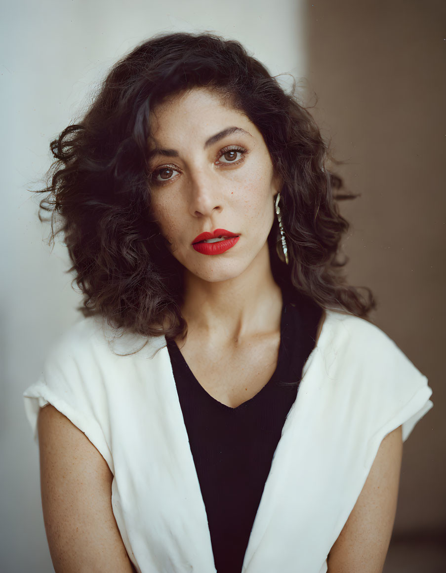 Woman with curly hair and red lipstick in white vest over black top gazes at camera.