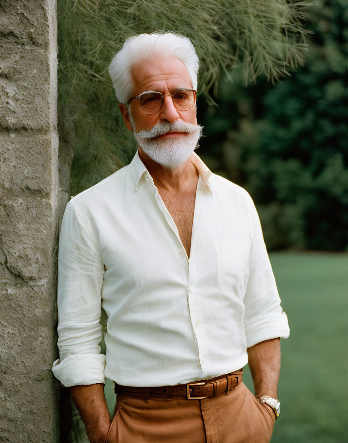 Elderly man with white hair and beard in glasses, white shirt, brown belt against greenery