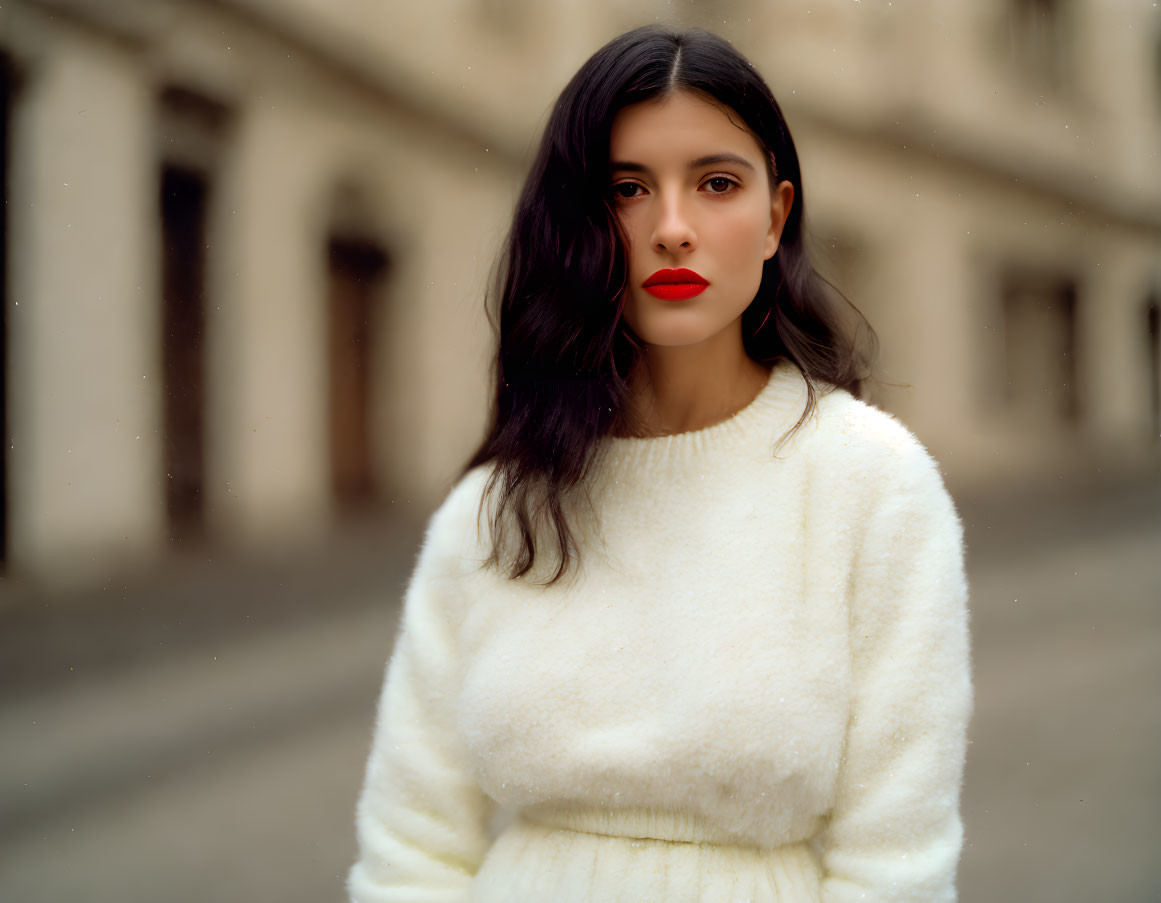 Dark-haired woman in red lipstick wearing white sweater in front of blurred building.