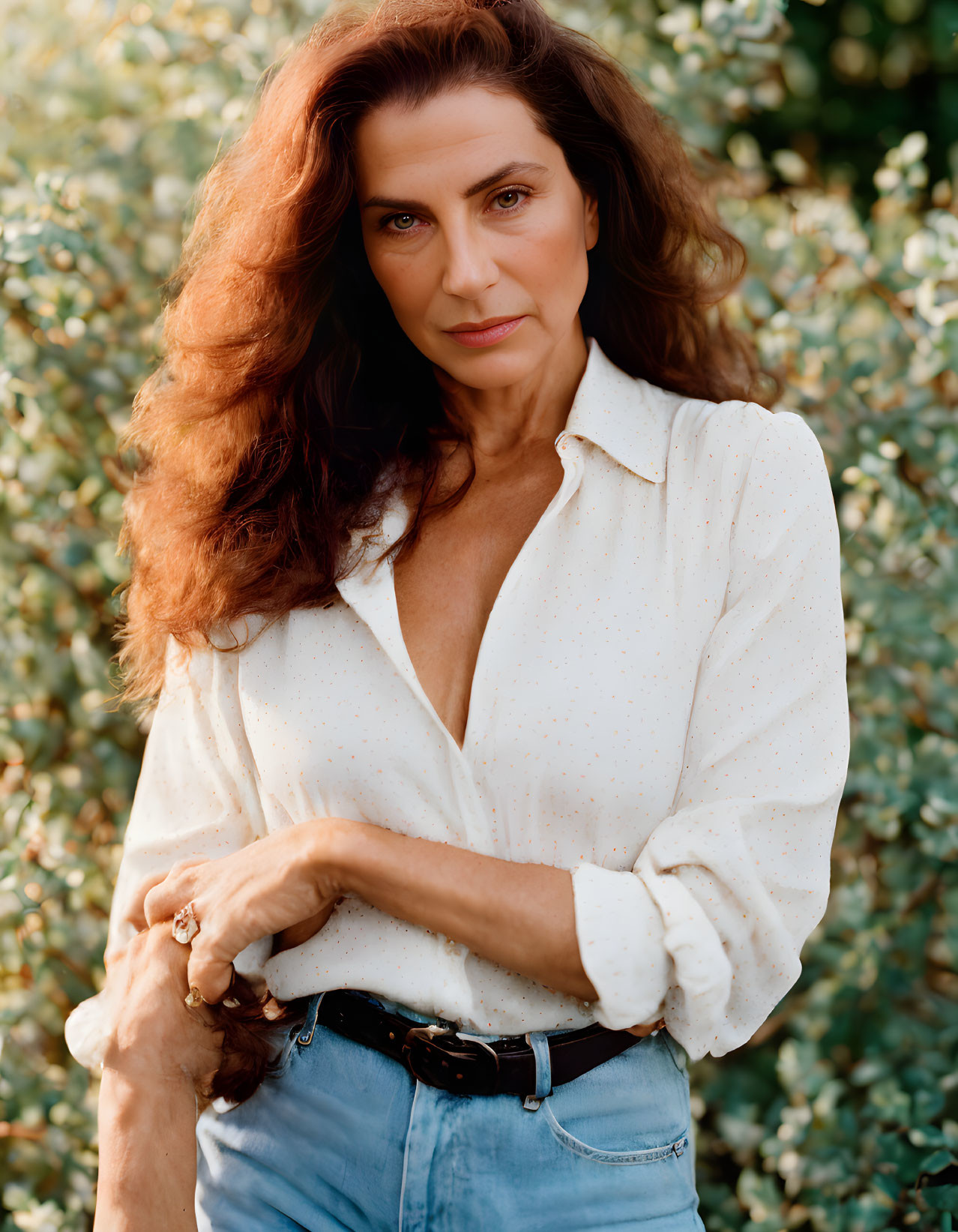 Long Curly-Haired Woman in White Dotted Shirt Poses in Green Setting