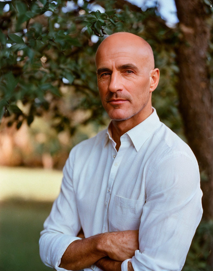 Stern bald man in white shirt with crossed arms by leafy tree