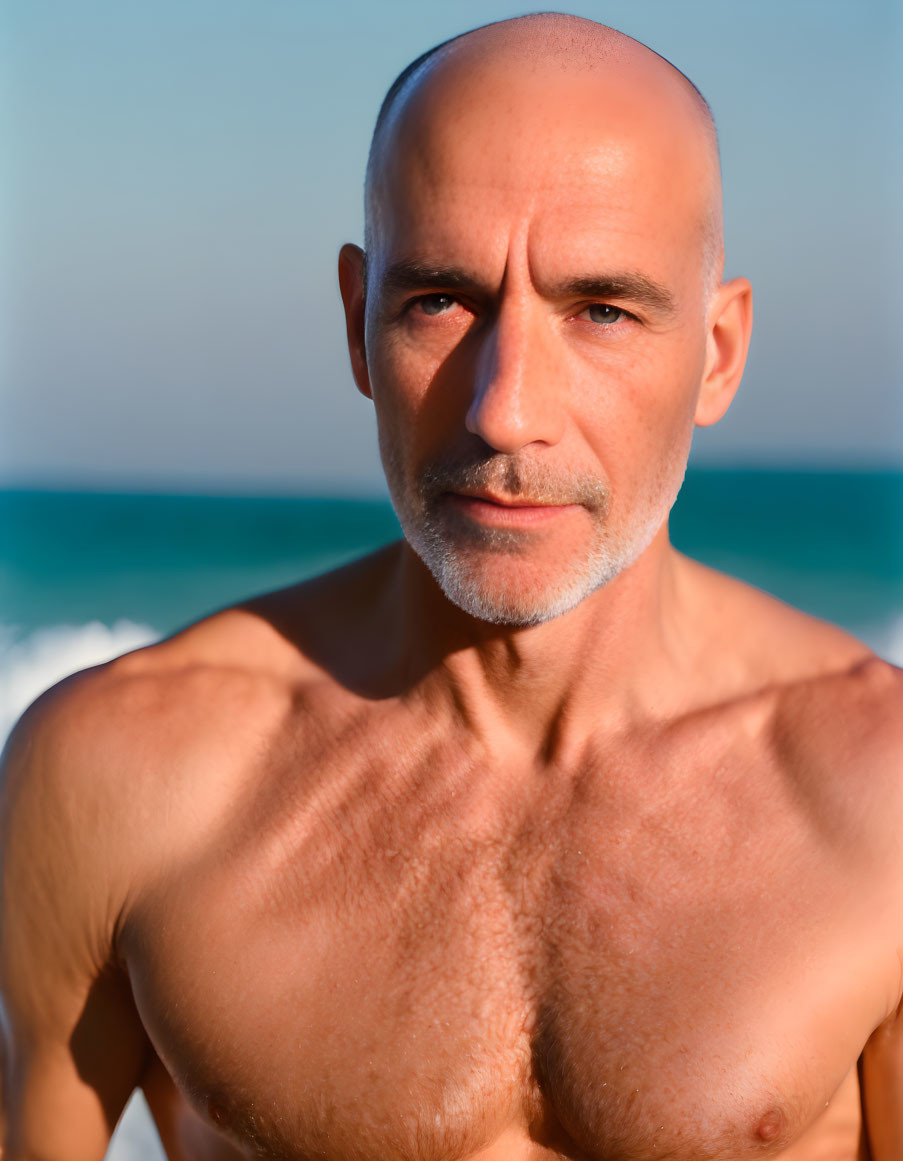 Bald man with beard on beach, shirtless and looking at camera