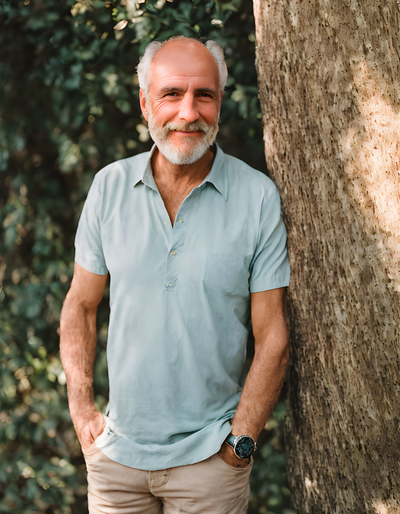 Elderly man in light blue shirt and beige pants leaning against tree