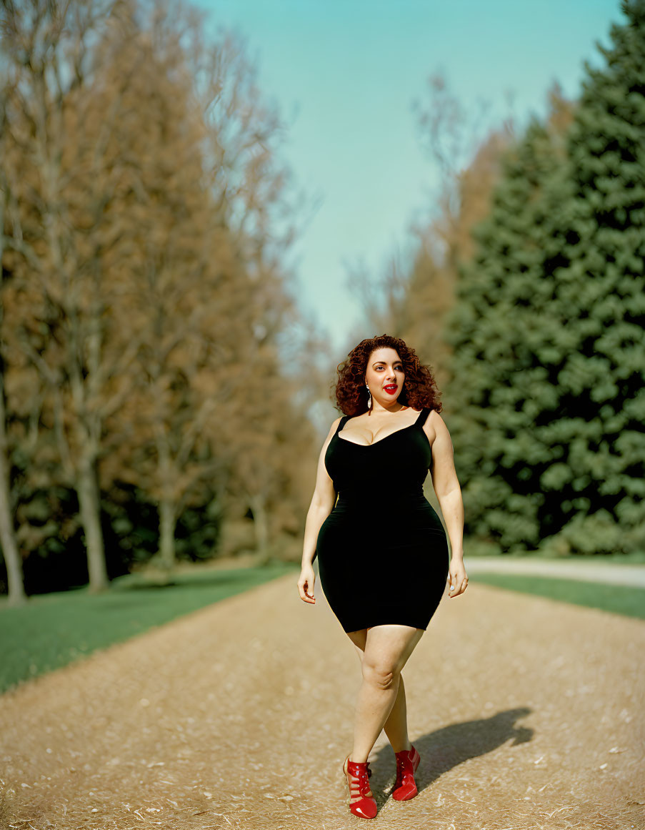 Woman in black dress and red boots walking on tree-lined path under clear blue sky