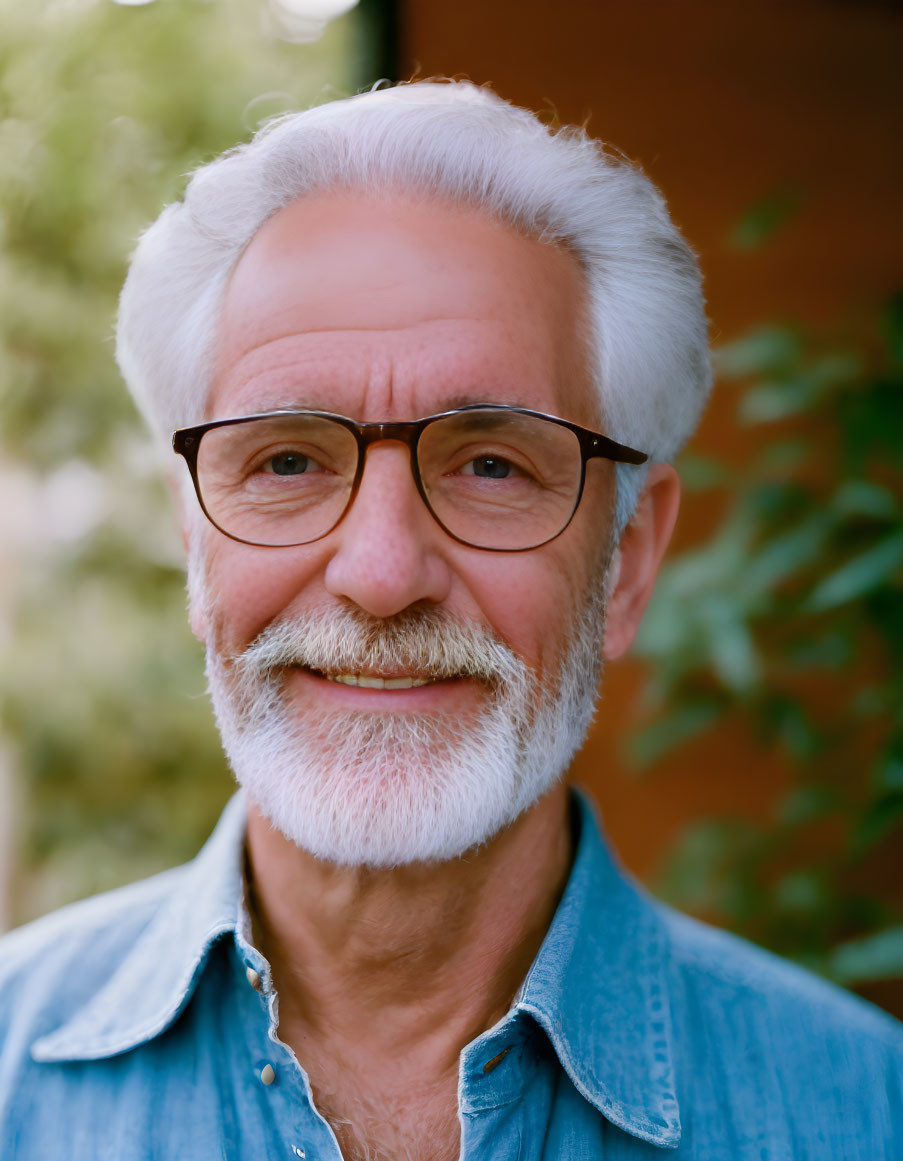 Elderly man with white beard and glasses in denim shirt smiling