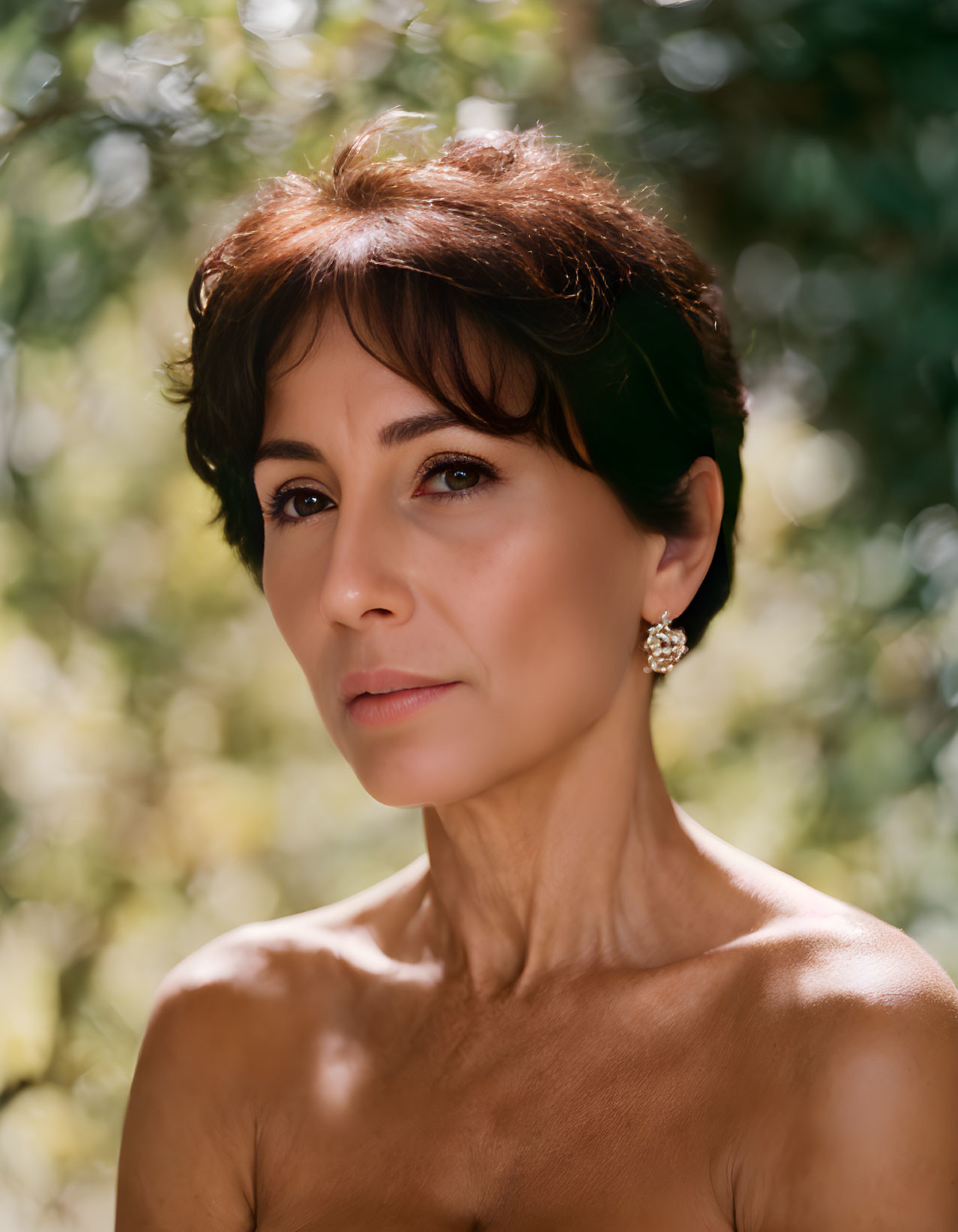 Short-haired woman with expressive eyes and dazzling earrings in front of greenery.