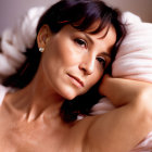 Dark-haired woman with earrings resting head on arm against pillow in soft-focus background