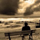 Person sitting on bench by stormy sea under dramatic sky with sun rays.