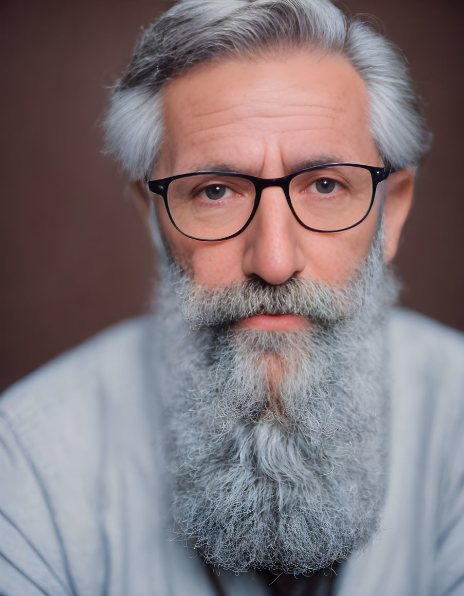 Gray-bearded elderly man in blue top and glasses staring at camera