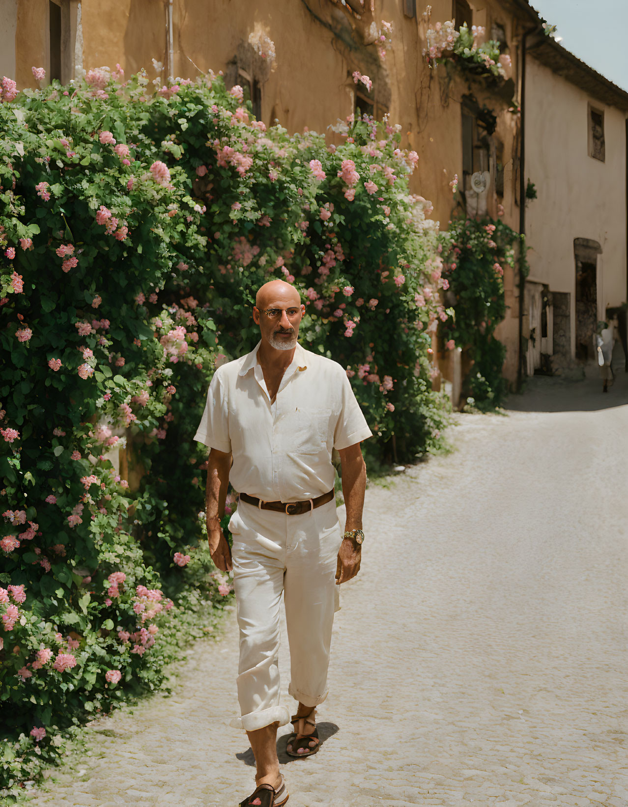 Man in white shirt strolling on cobblestone street with pink flowers