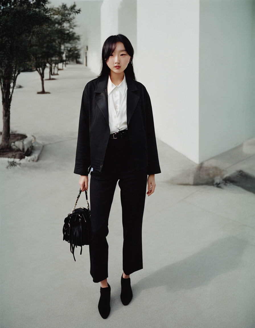 Woman in black blazer and trousers with white blouse and ankle boots holding fringed handbag