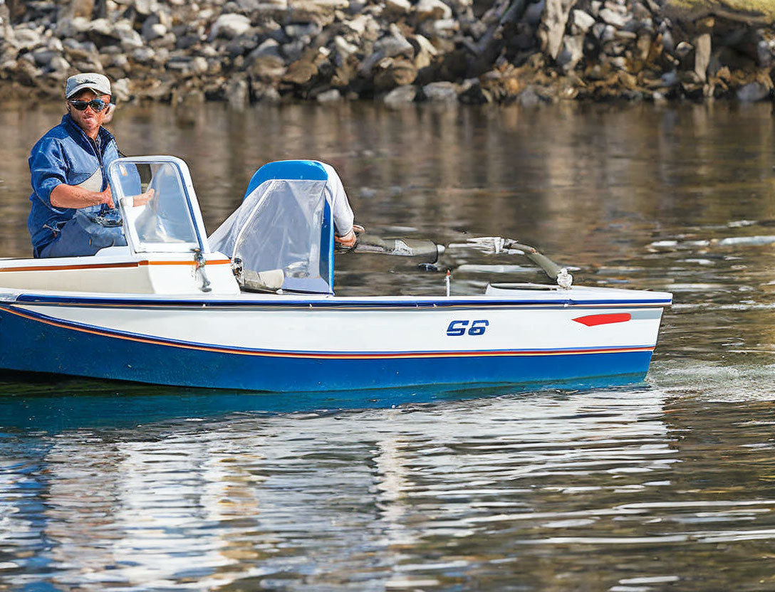 Man in sunglasses and cap steering white and blue motorboat near rocky shore