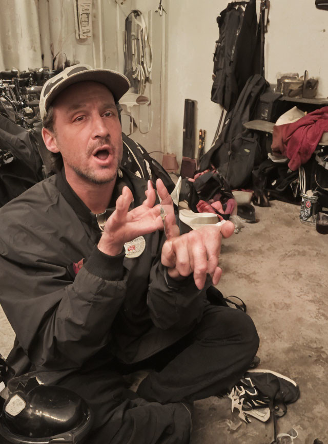 Man in cap and overalls gesturing in room with tools.