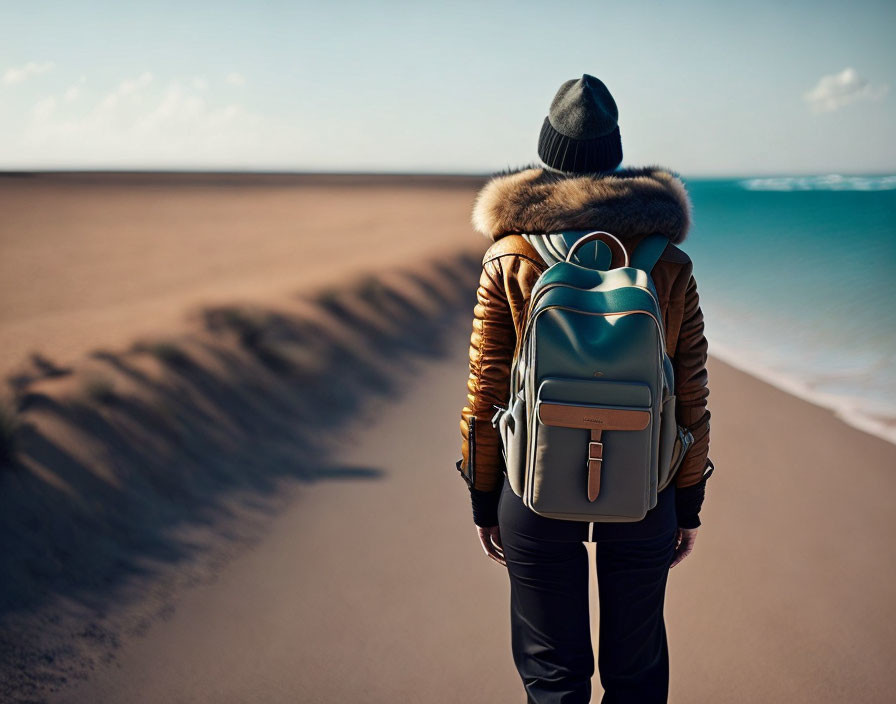 Person in winter jacket and beanie at sandy beach facing ocean