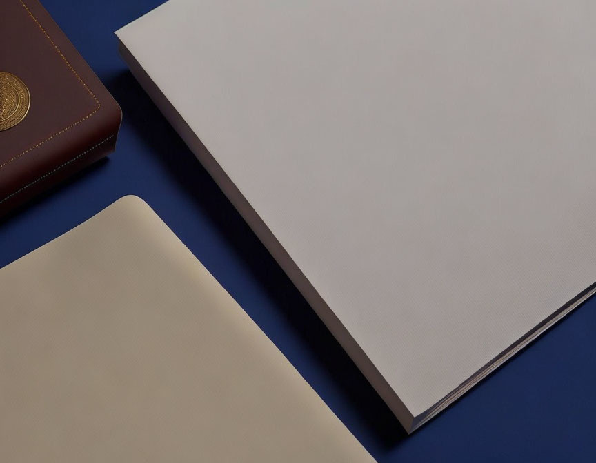 Closed brown notebook, white paper, and open beige folder on dark blue surface