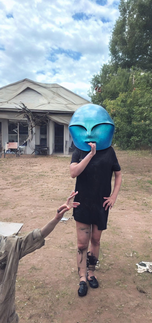 Person wearing oversized alien head mask in front of dilapidated building with reaching hand