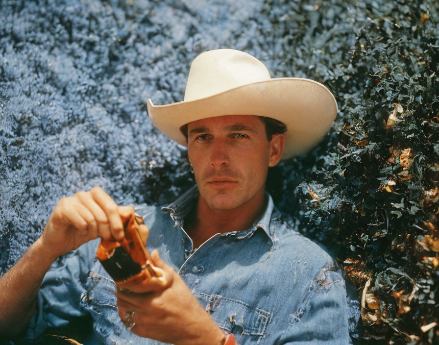 Cowboy man in denim shirt with hat sitting against blue shrubbery holding object