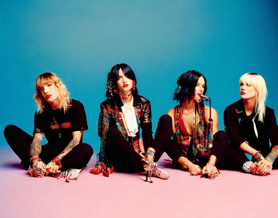 Four stylish women in black with colorful scarves and tattoos against blue backdrop