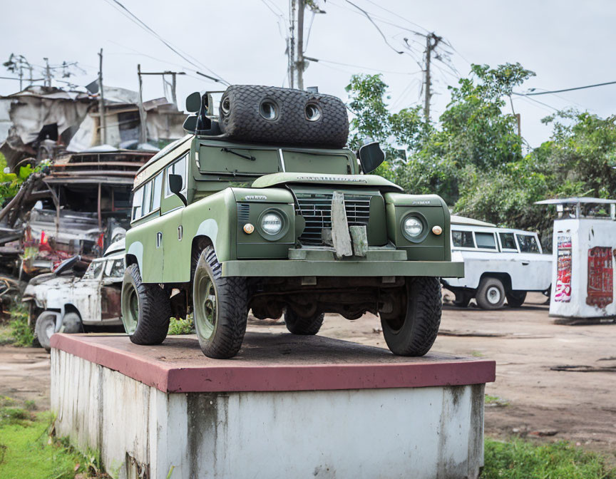 Vintage military-style green vehicle on concrete pedestal in yard with cars.