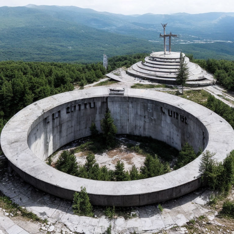 Dilapidated circular concrete structure with grand cross and tower on hilltop surrounded by forested landscape