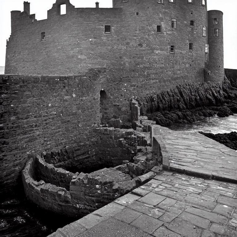 Grayscale image of rugged stone castle by the sea with aged walls and small boat dock under overcast