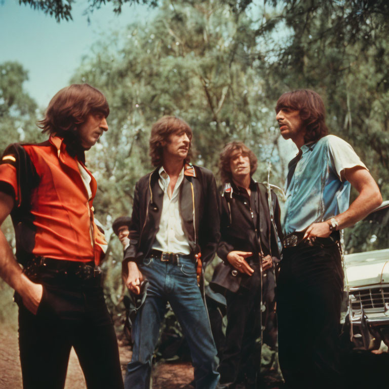 Four men in retro clothing converse outdoors with vintage car and trees.