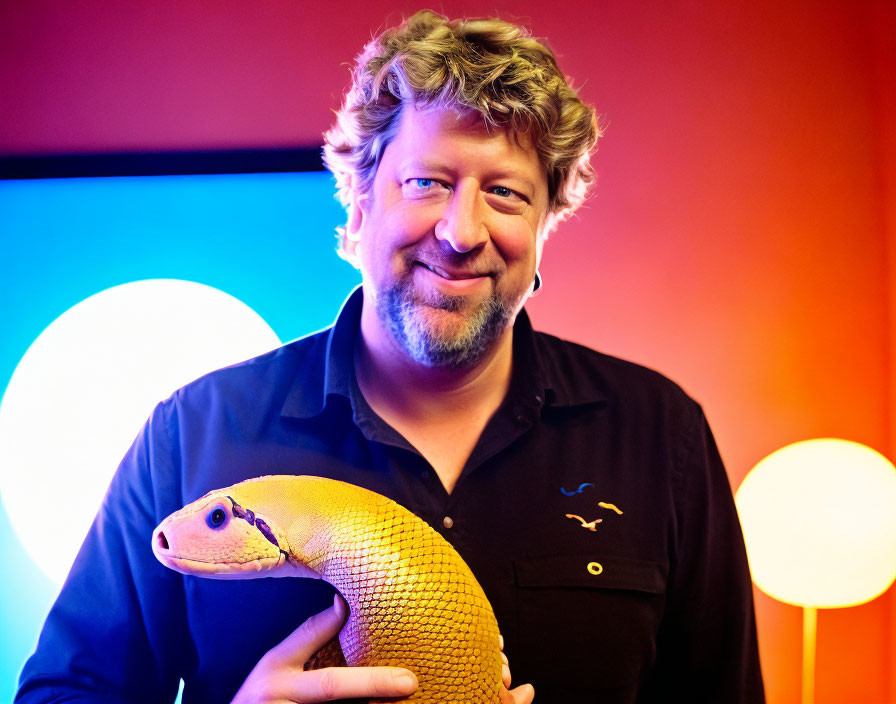 Smiling person with curly hair holding a yellow lizard replica amid colorful lights