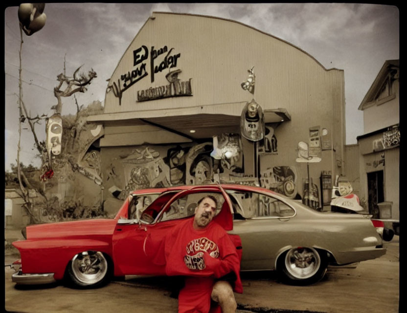 Man in red shirt leaning on classic red car in front of eclectic building with "El Paso Wre