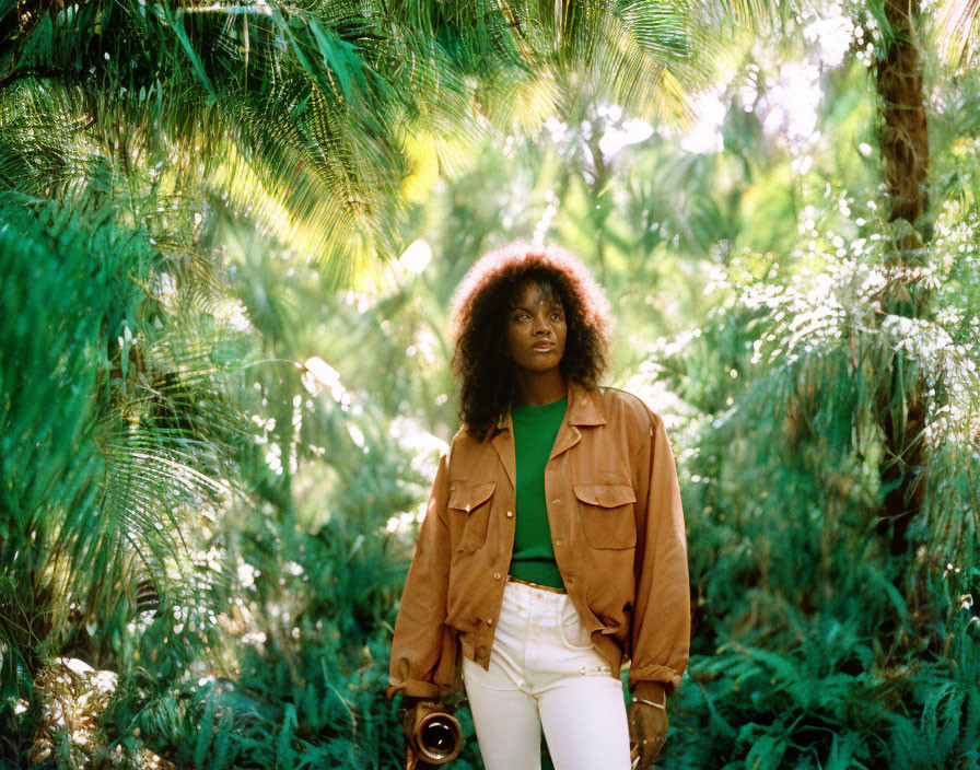 Woman with afro hairstyle in brown jacket and white pants among lush green foliage with camera