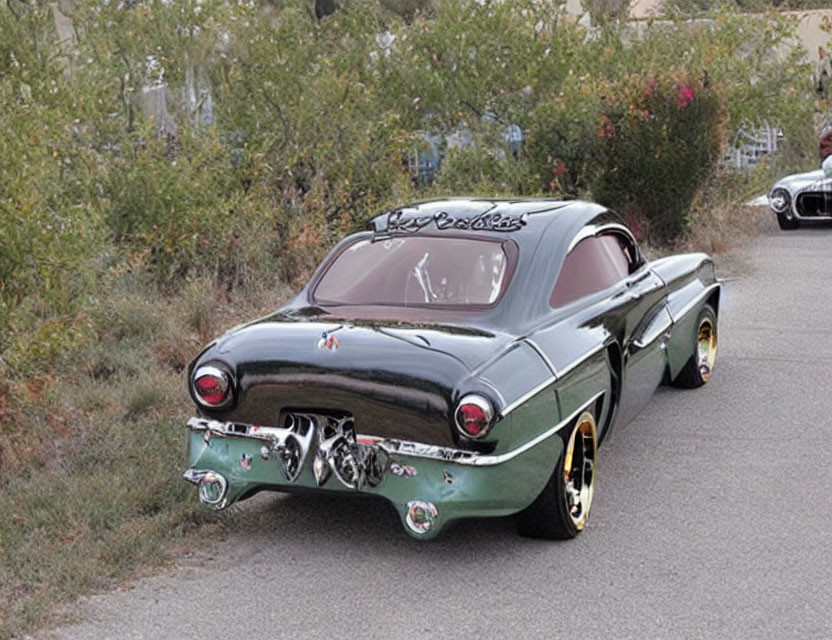 Vintage green car with flame decals and white wall tires parked by roadside shrubs.