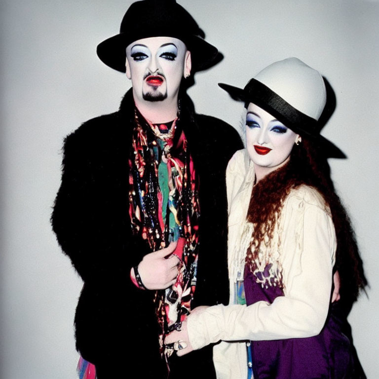 Two individuals in dramatic makeup and artistic attire embracing in hats and scarves