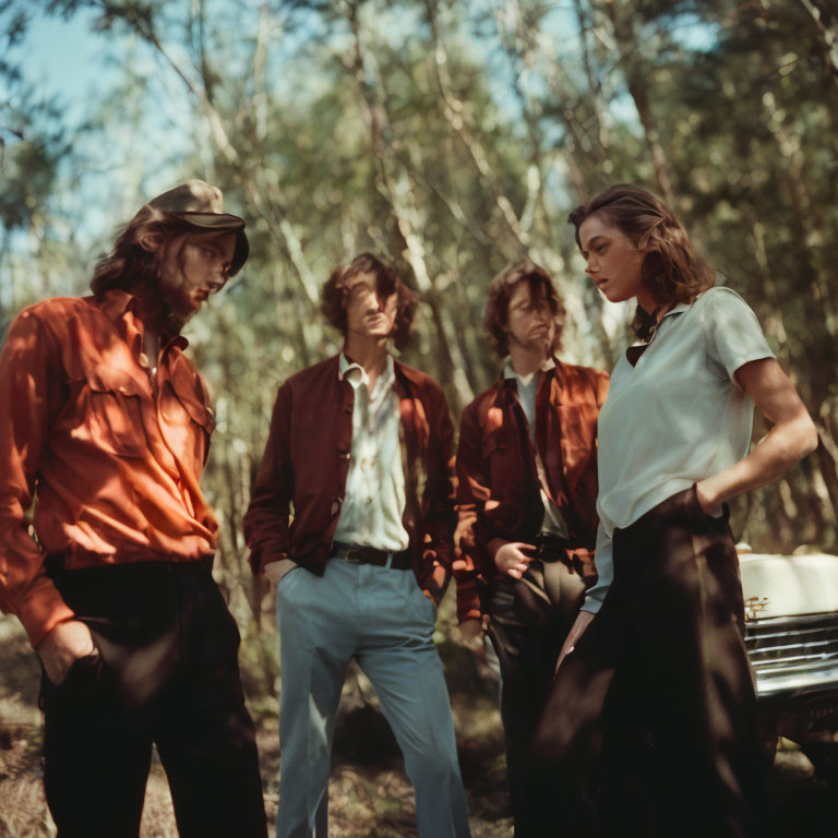 Vintage attired group near old car in wooded area.