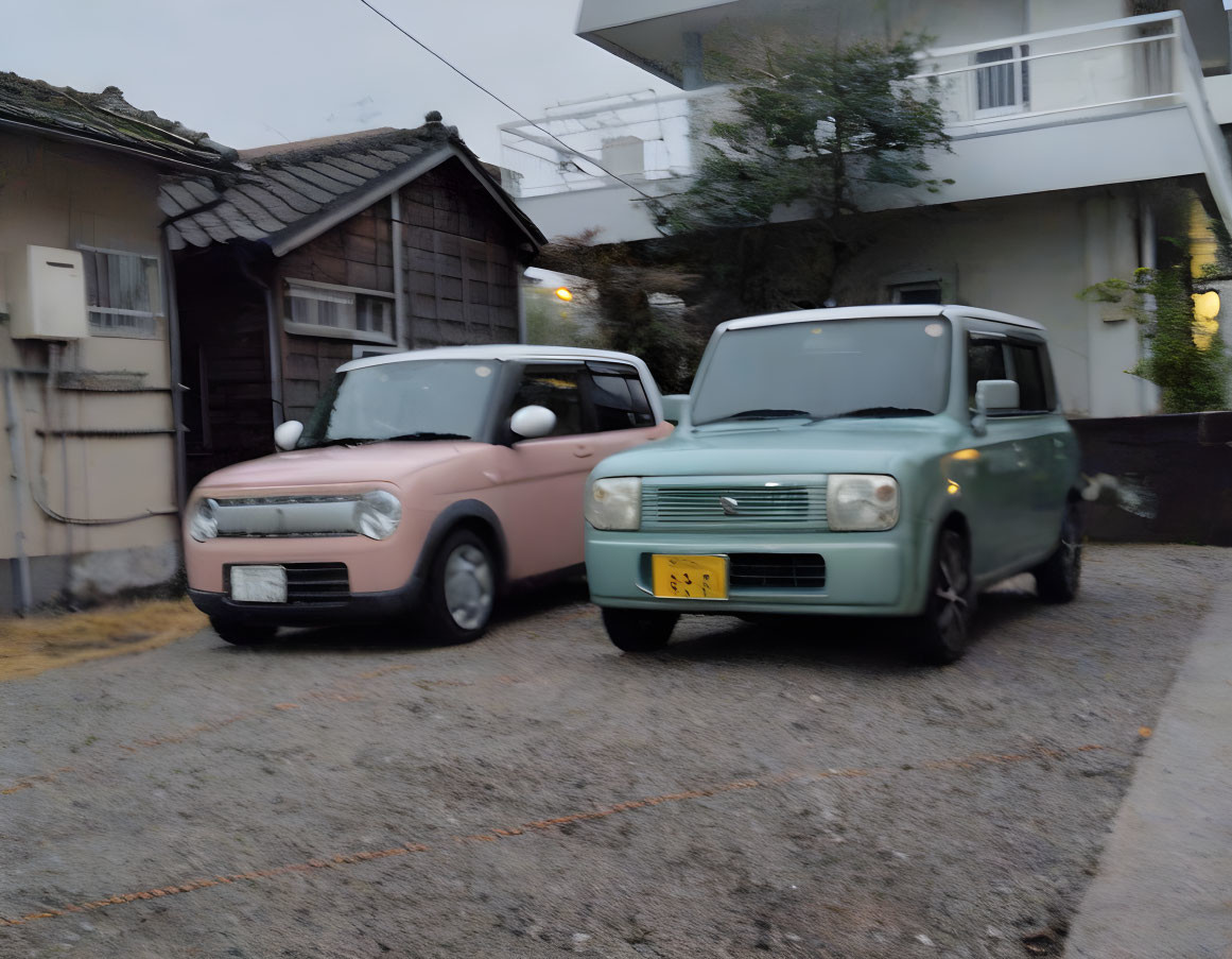 Compact pink and green cars parked by wooden house and concrete building.