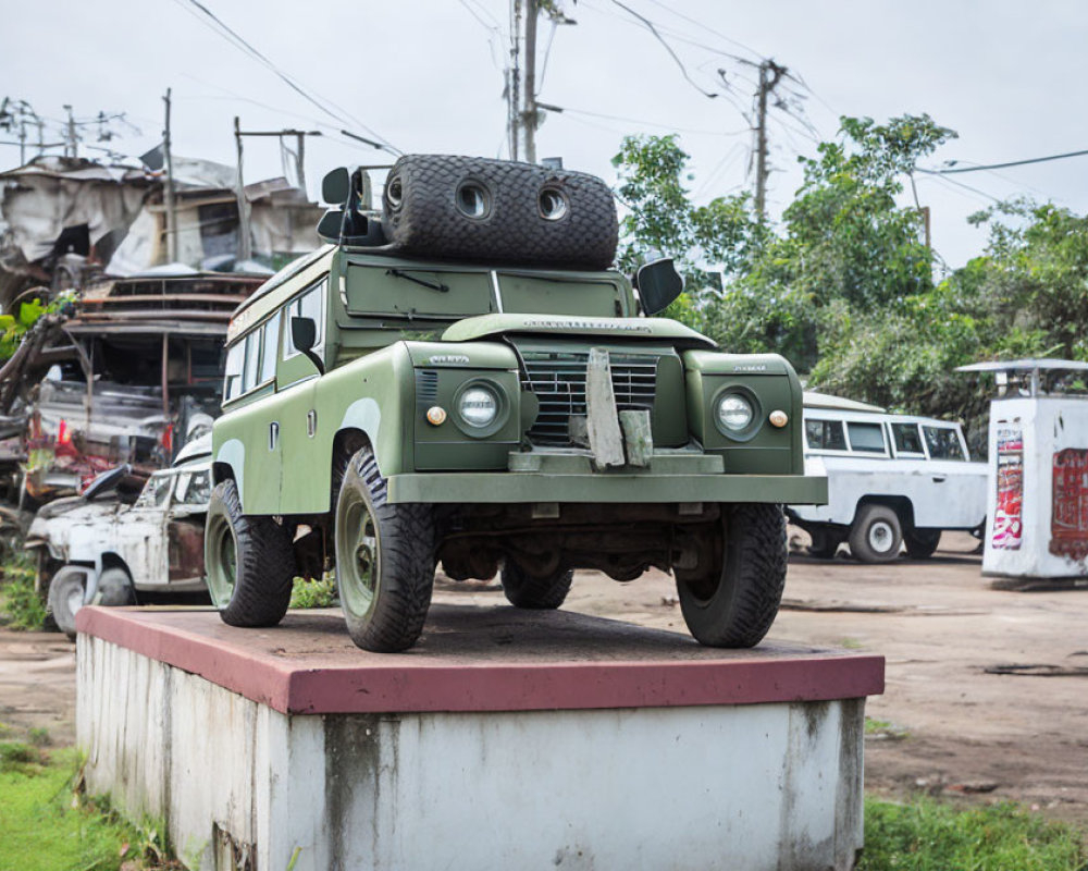 Vintage military-style green vehicle on concrete pedestal in yard with cars.