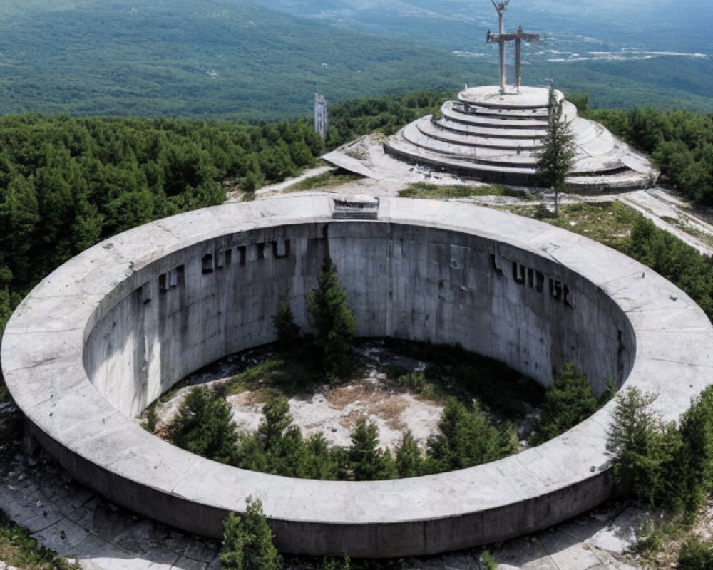 Dilapidated circular concrete structure with grand cross and tower on hilltop surrounded by forested landscape