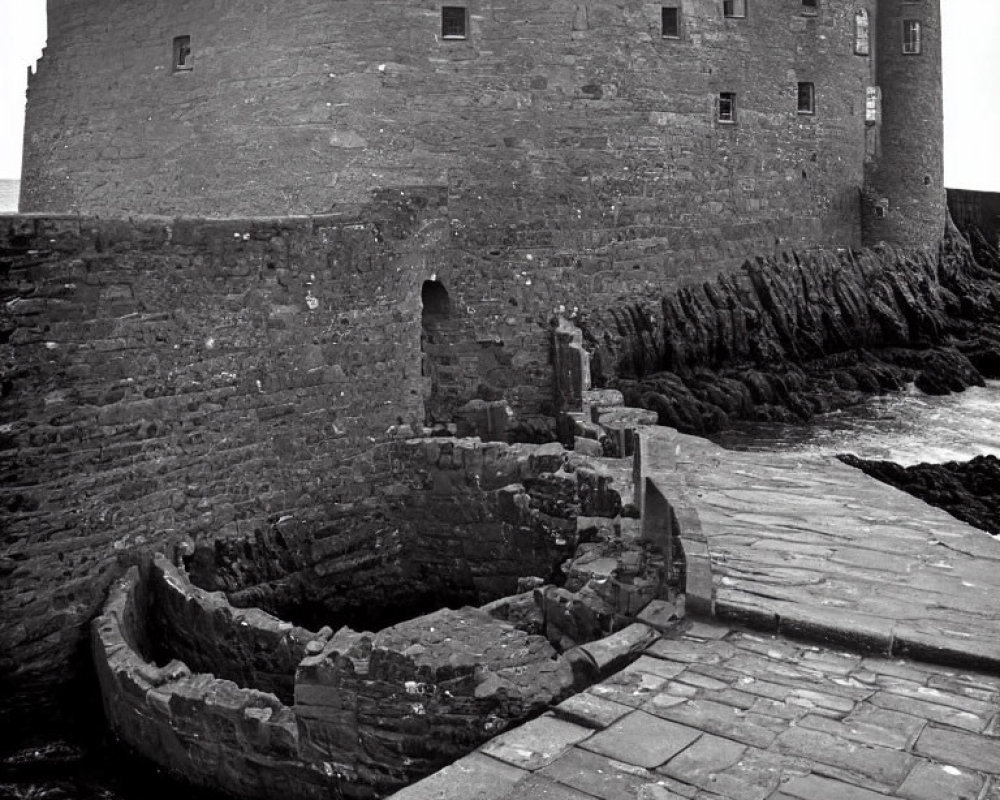 Grayscale image of rugged stone castle by the sea with aged walls and small boat dock under overcast