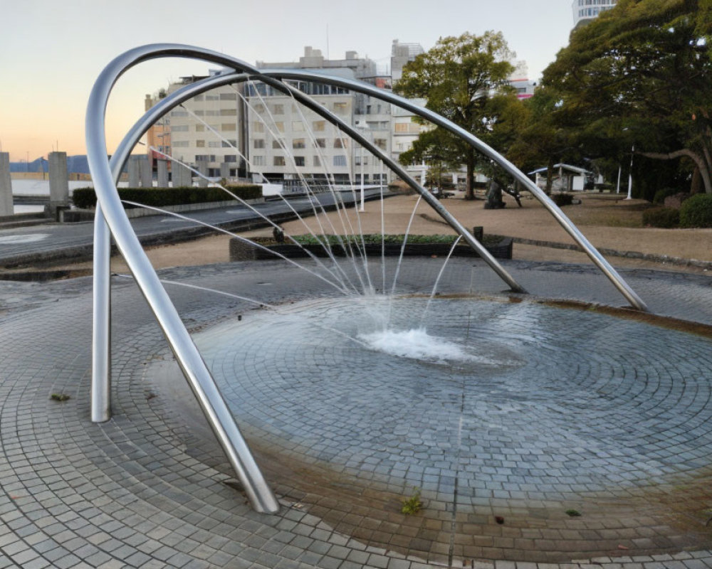 Urban fountain with arched metal structures and circular water pool amid buildings and trees.