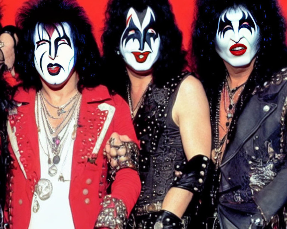 Three individuals in flamboyant rock attire and iconic makeup on a red background.