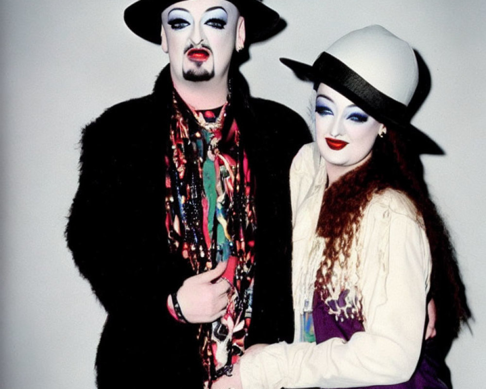 Two individuals in dramatic makeup and artistic attire embracing in hats and scarves