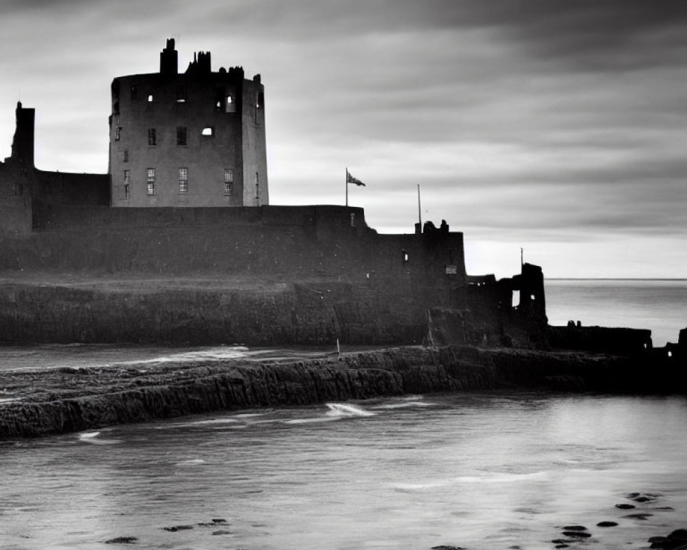 Monochrome image: ancient castle by the sea with flag, calm water, dramatic sky