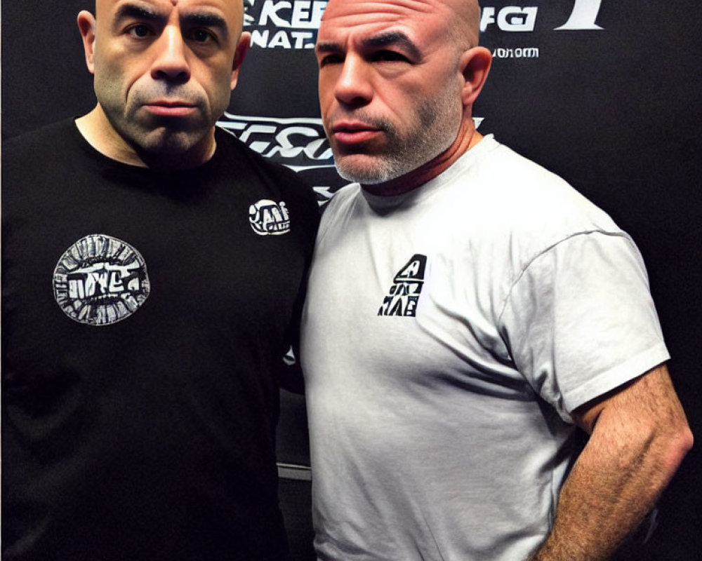 Two bald men in black t-shirts with logos, one with a beard, posing in front of logo