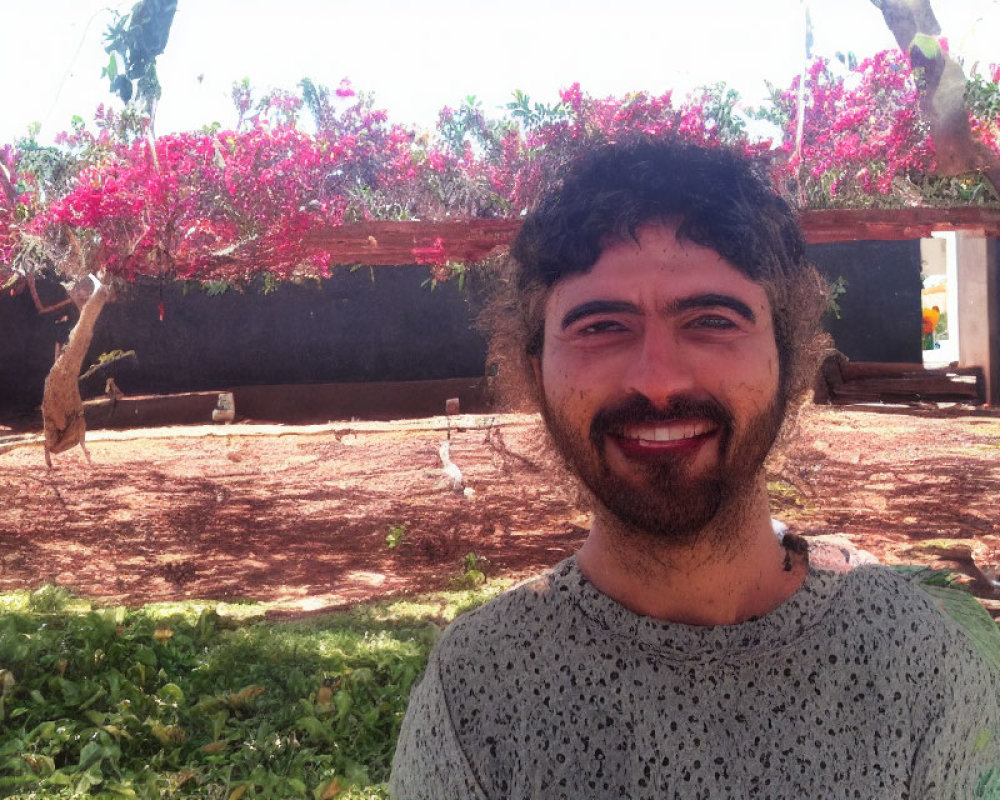 Bearded man smiling outdoors with pink flowers and wooden trellis