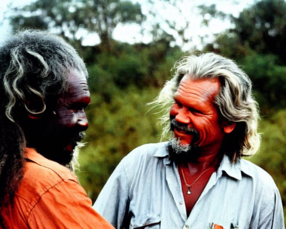 Men with painted faces in conversation: one in orange attire with traditional markings, the other in grey shirt