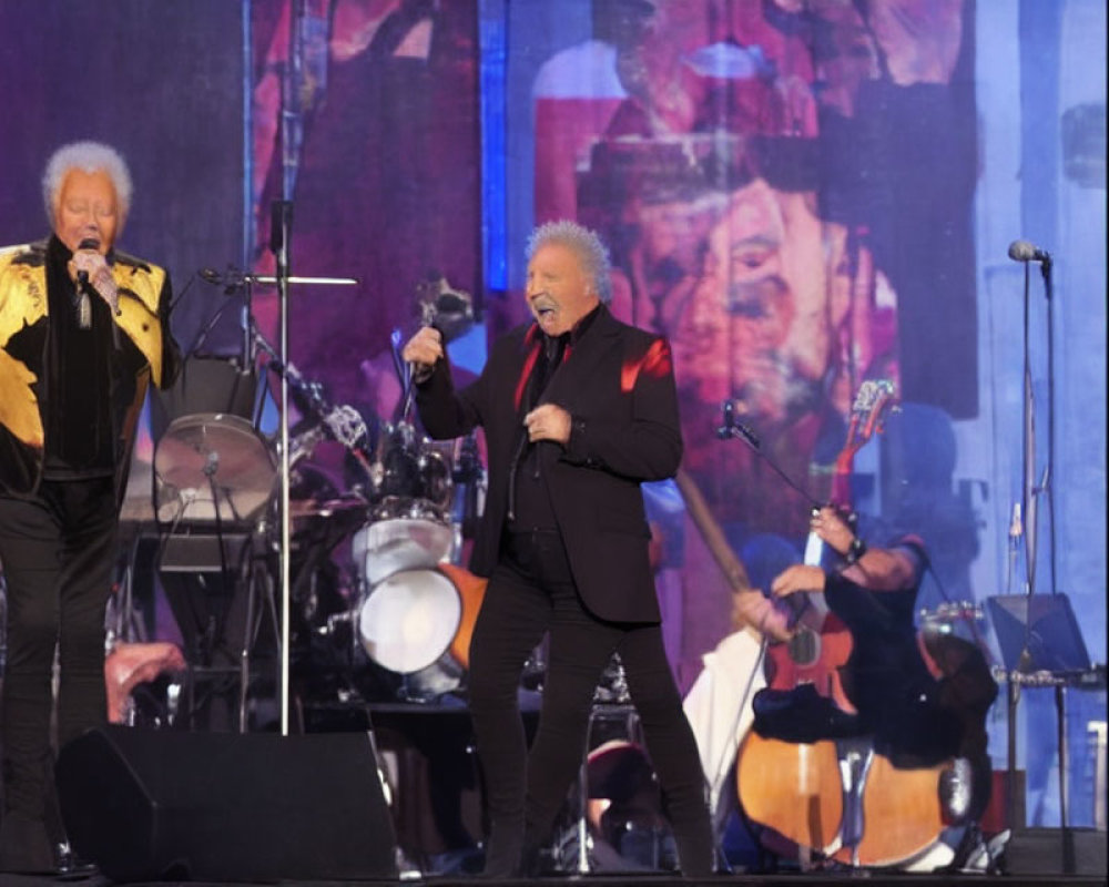 Two male singers in gold and black jackets perform on stage with a band against a backdrop showcasing their images