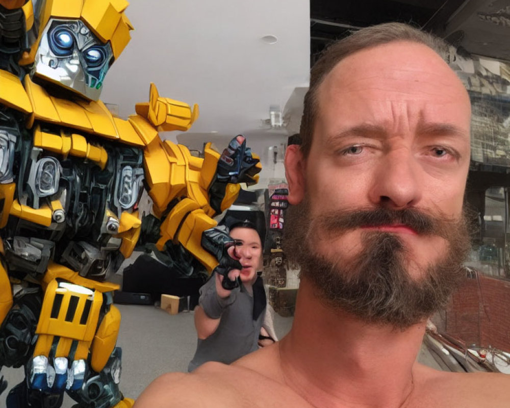 Bearded man takes selfie with Bumblebee Transformer and person aiming gun in background.