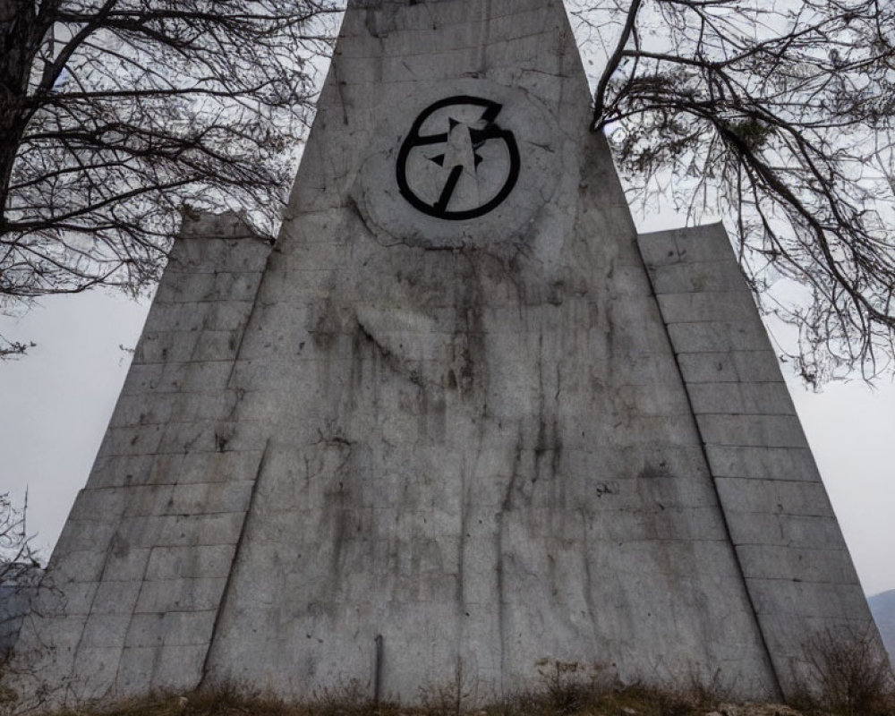 Weathered concrete monument with cyclist symbol and bare trees under cloudy sky